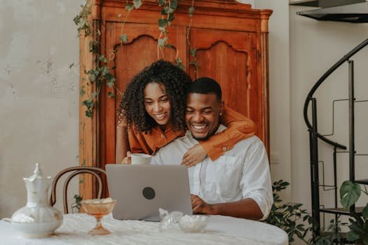 Smiling couple embracing while looking at a laptop in a cozy indoor setting, enjoying togetherness.