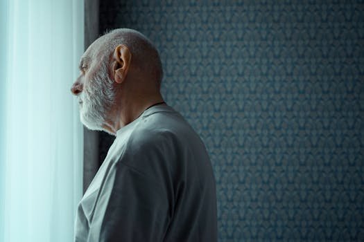 A senior man with a beard looks thoughtfully out of a sunny window, reflecting on life indoors.