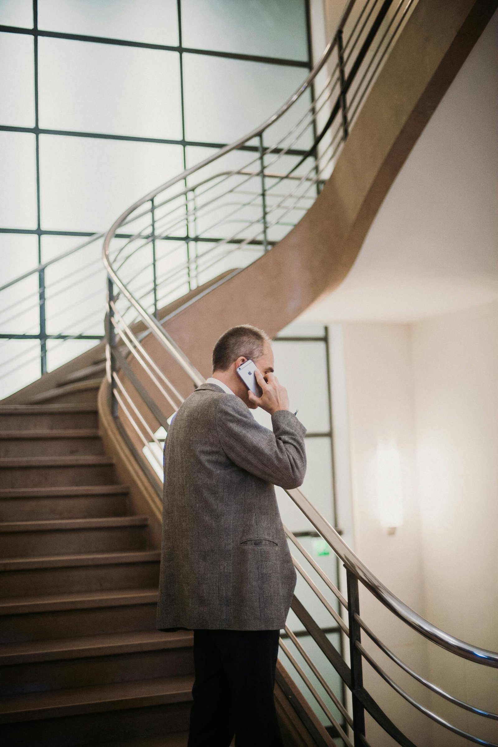 A businessman in a suit talks on his smartphone by a modern staircase indoors.